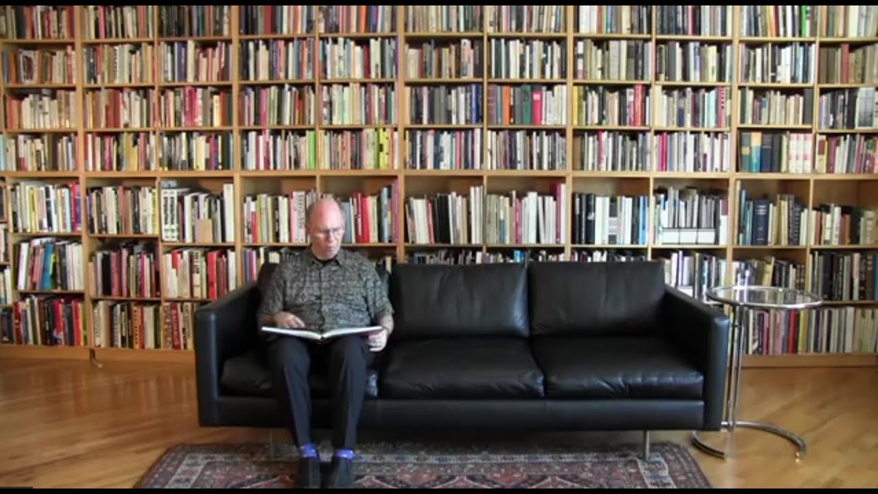 Victor Margolin, a bald older gentleman, sits in a scholarly pose reading a book on a black leather couch in front of a wall of bookshelving packed with several thousand books
