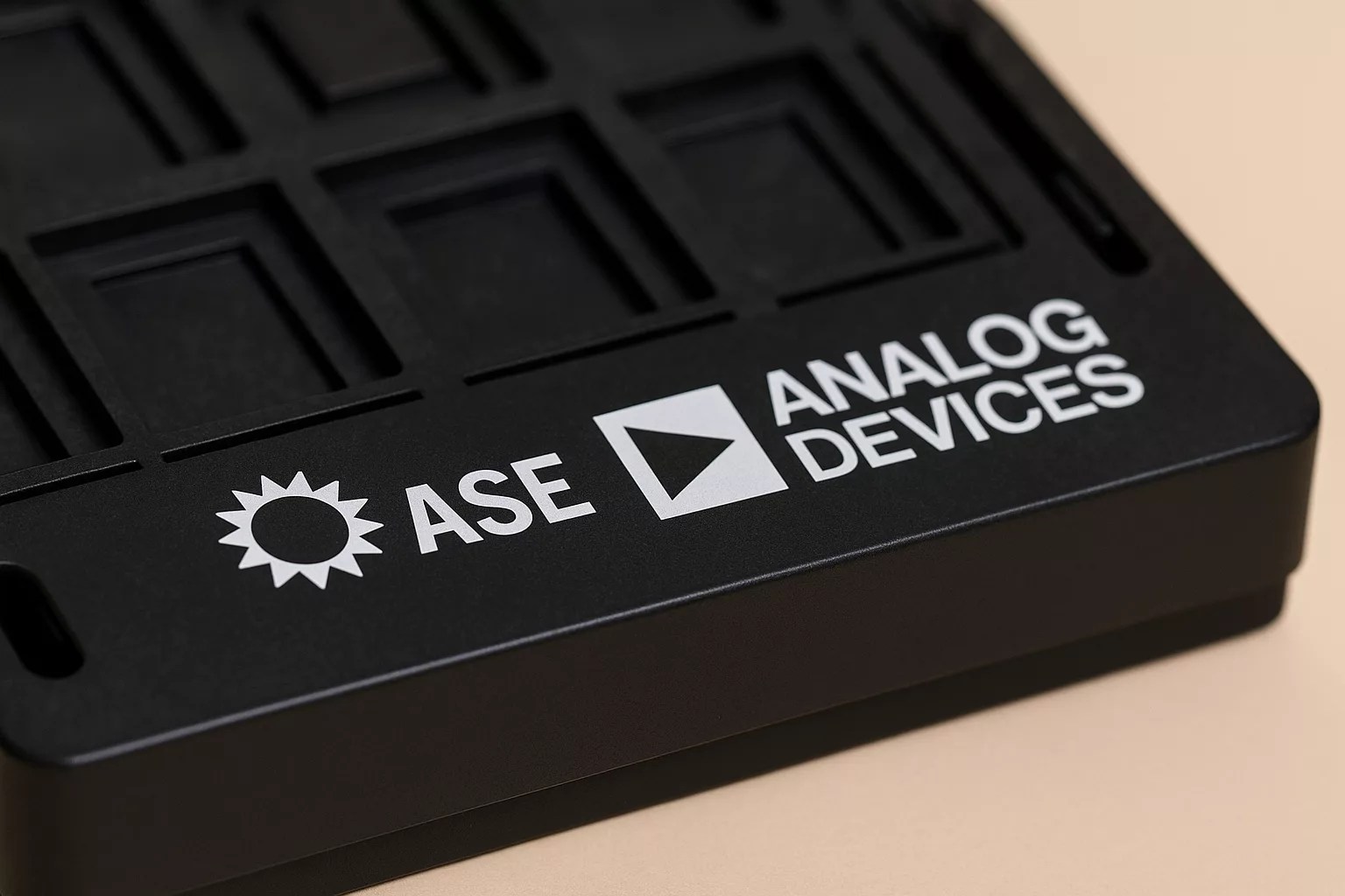 A close-up of a black plastic component tray, commonly used in OSAT facilities, with the ASE and Analog Devices logos and text printed in white on its surface.