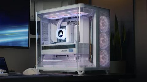 A white Corsair AIR 5400 gaming PC case with RGB lighting is displayed on a desk. Featuring triple-chamber airflow, its clear side panels reveal cooling fans and neatly arranged components, with a monitor and plant in the background.