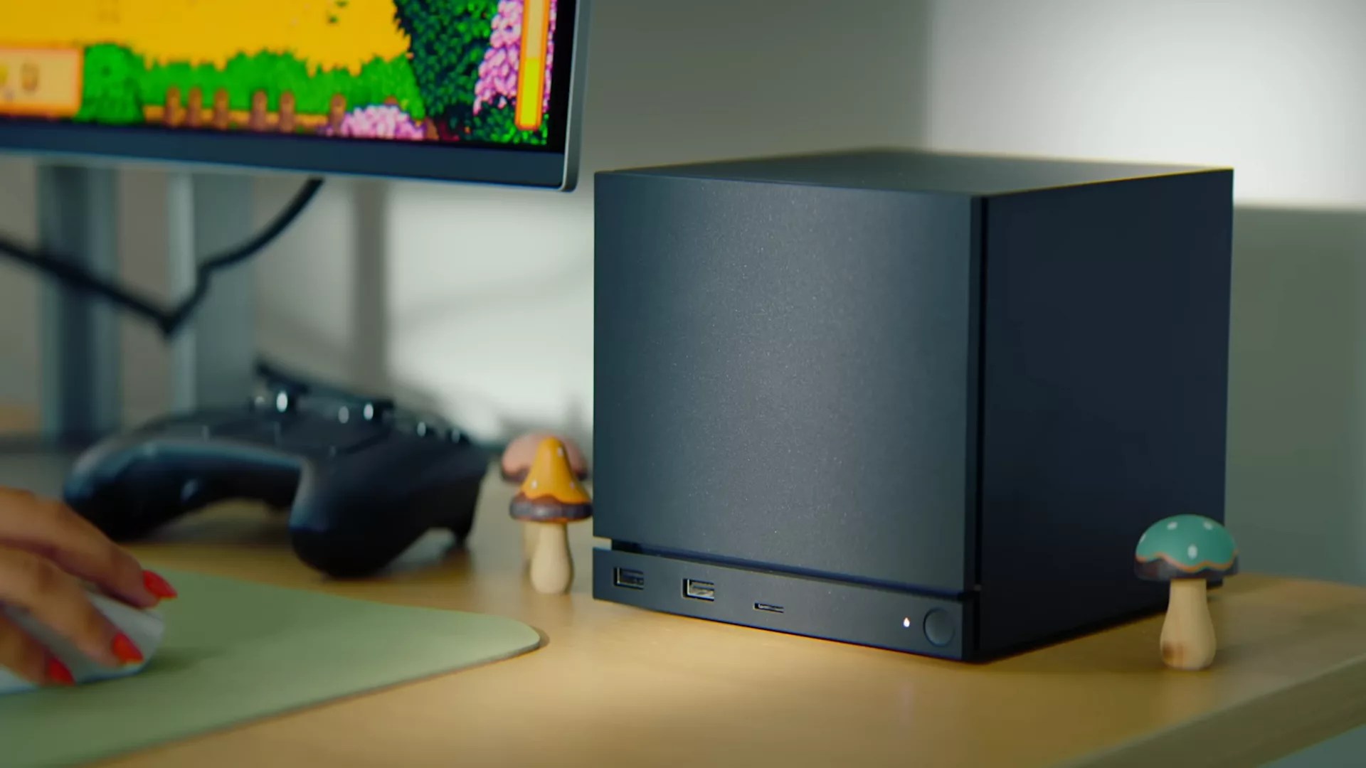 A black, cube-shaped mini PC resembling a Steam Machine sits on a desk near a monitor, gaming controller, and two small decorative mushroom figures. A hand with painted nails uses a mousepad as the monitor displays a colorful pixel art game.