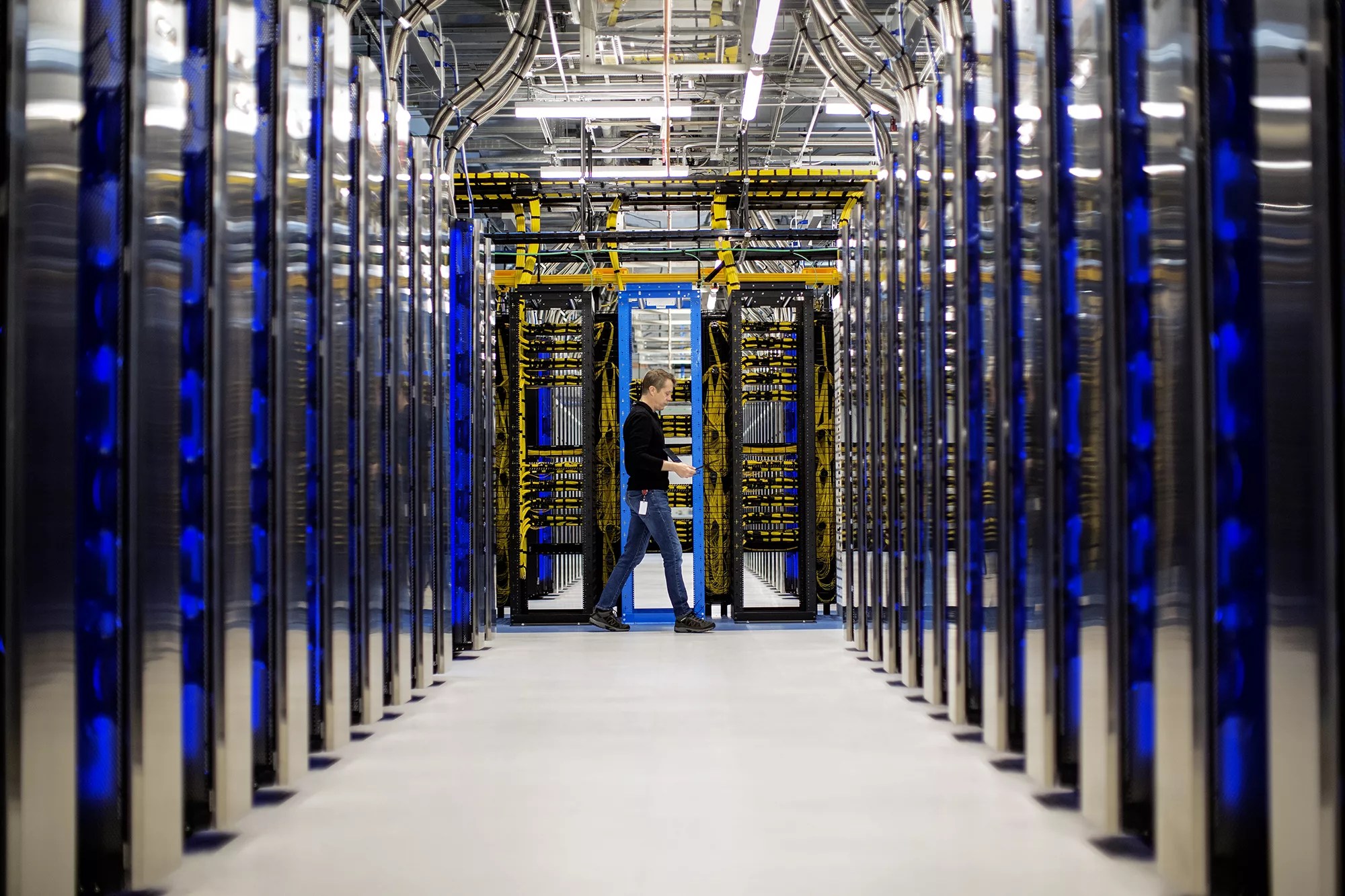 A person walks through a brightly lit Fairwater Datacenters aisle lined with rows of tall server racks filled with blue and yellow cables and equipment.