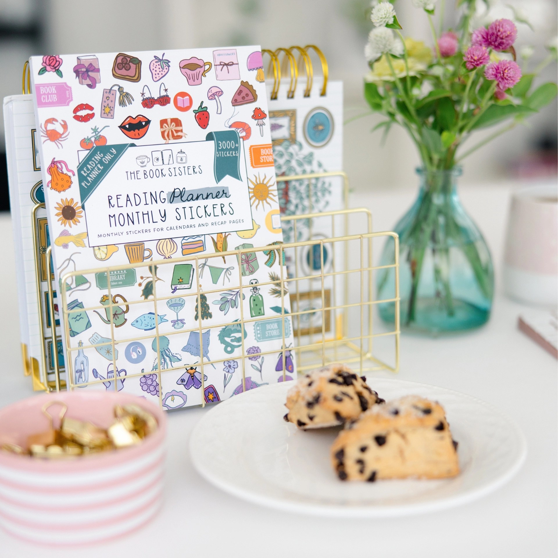 The Book Sisters Reading Planner Monthly Stickers cover sheet with 3000+ illustrated stickers including books, food, flowers, and cozy icons propped up in book stand with scones, nuts and flowers nearby