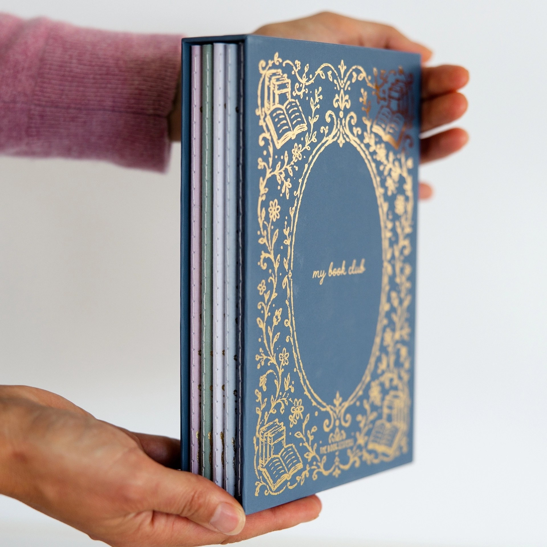 Two hands holding the navy My Book Club journal box with five colorful fanned journals inside.