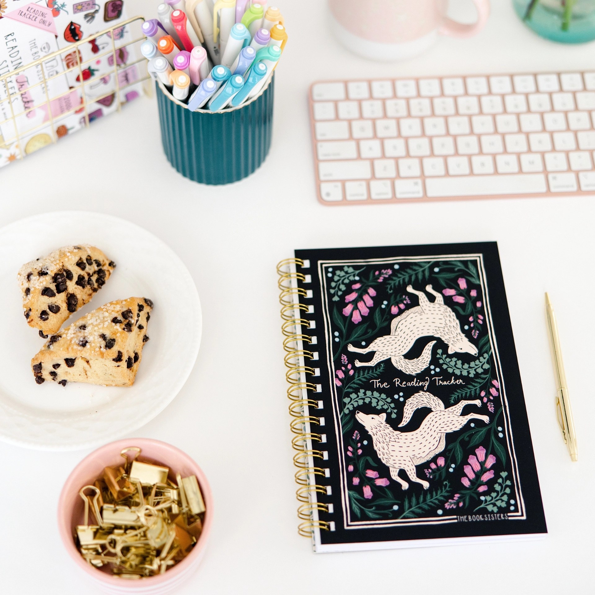 The Reading Tracker notebook styled on a desk with colorful pens, a scone, gold binder clips, and a keyboard
