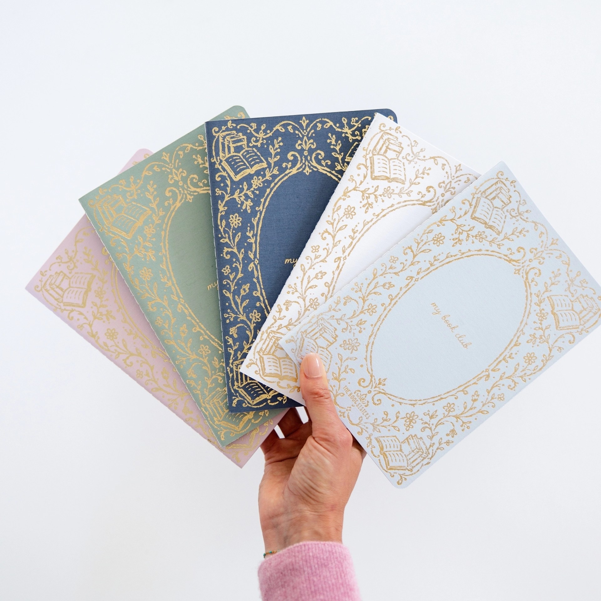 Hand fanning out five My Book Club journals in pink, sage, navy, cream, and light blue covers.