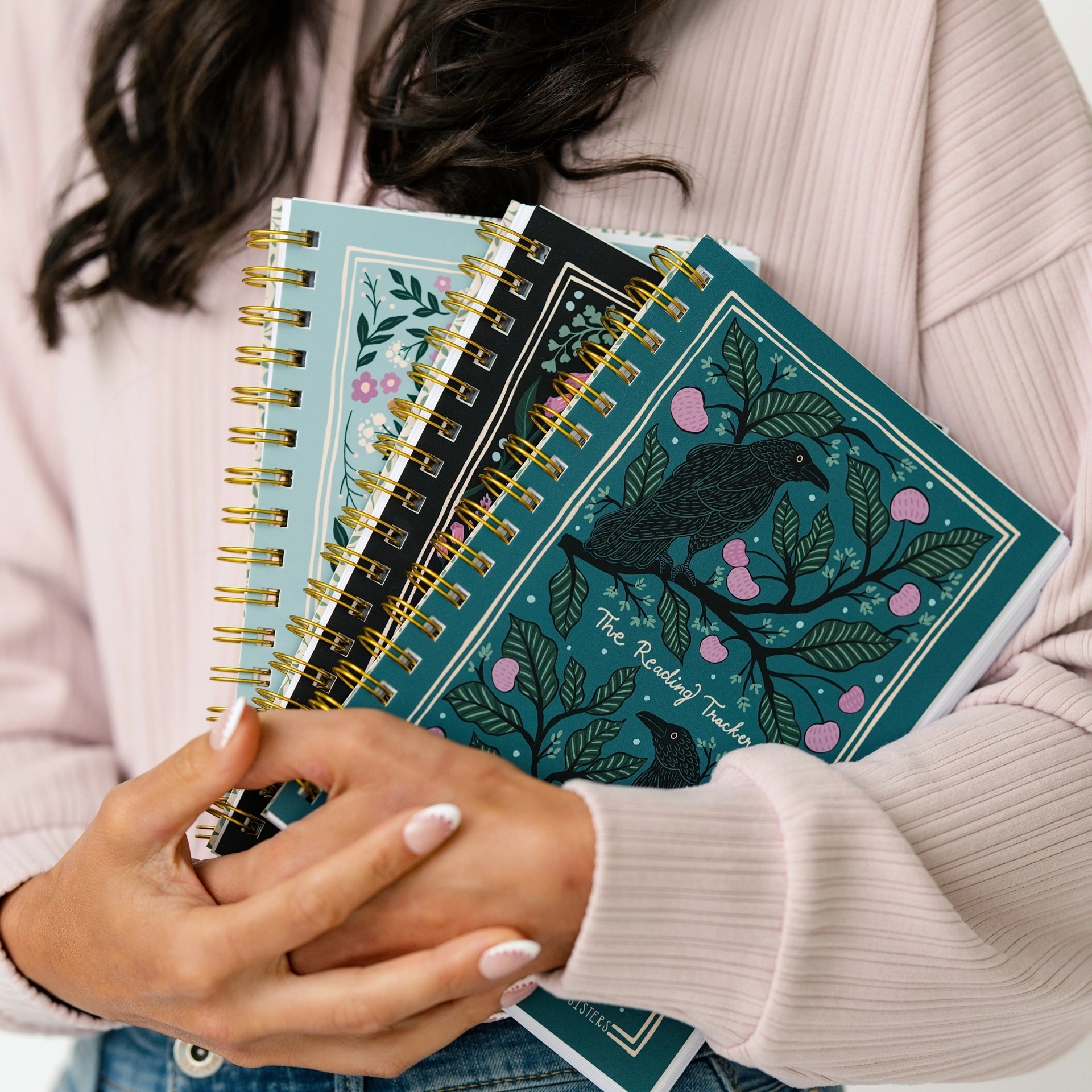 Three Reading Tracker notebook covers fanned out and held, showing the teal raven, black fox, and light blue cat designs