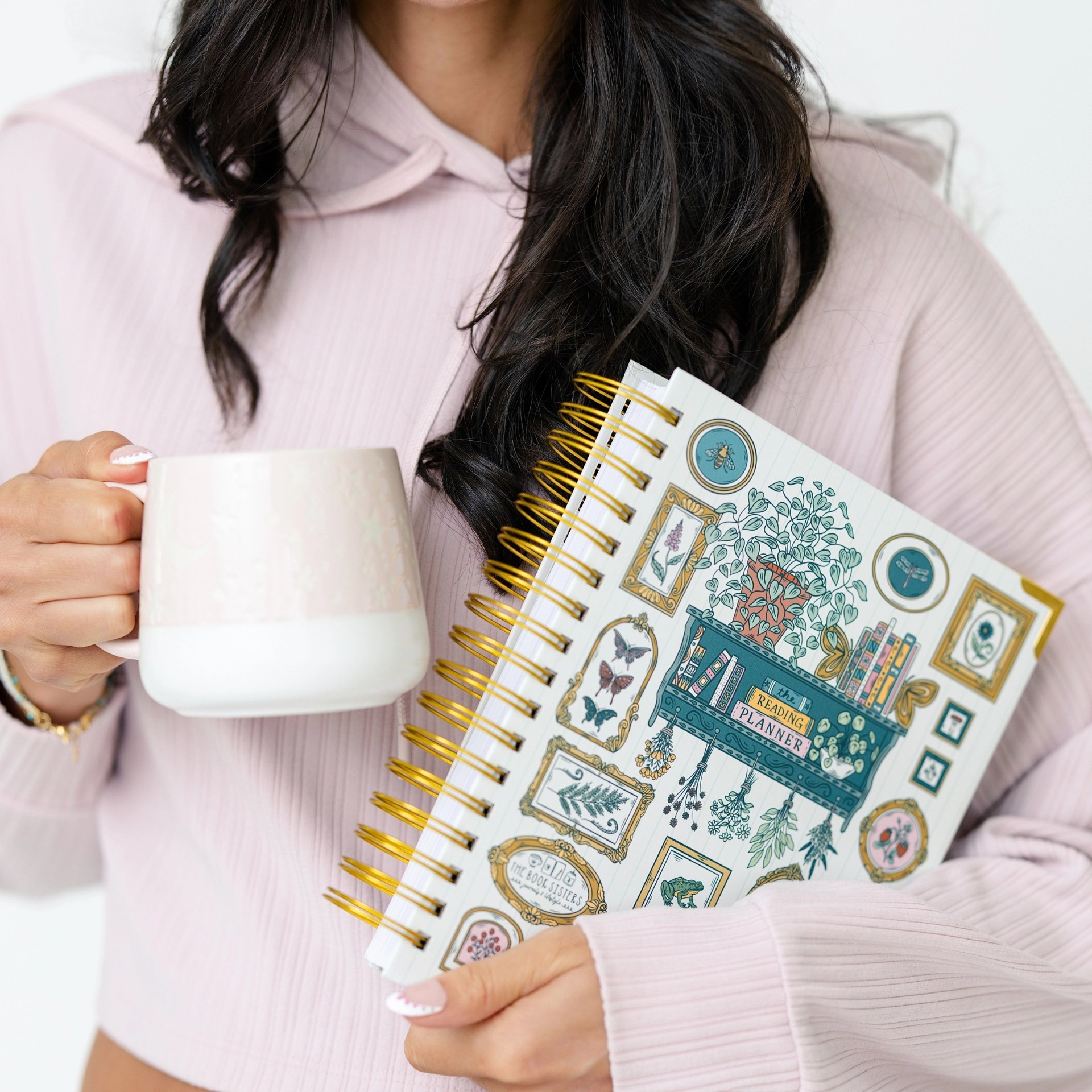 Woman in a pink sweater holding the garden-themed reading planner and a latte, close-up on the gold wire binding