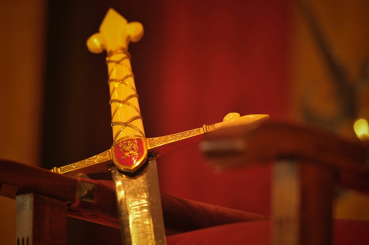 a close up of the hilt of a sword hanging on some sort of chair armrest. The hilt is yellow with gold accents and it's on top of a red background.