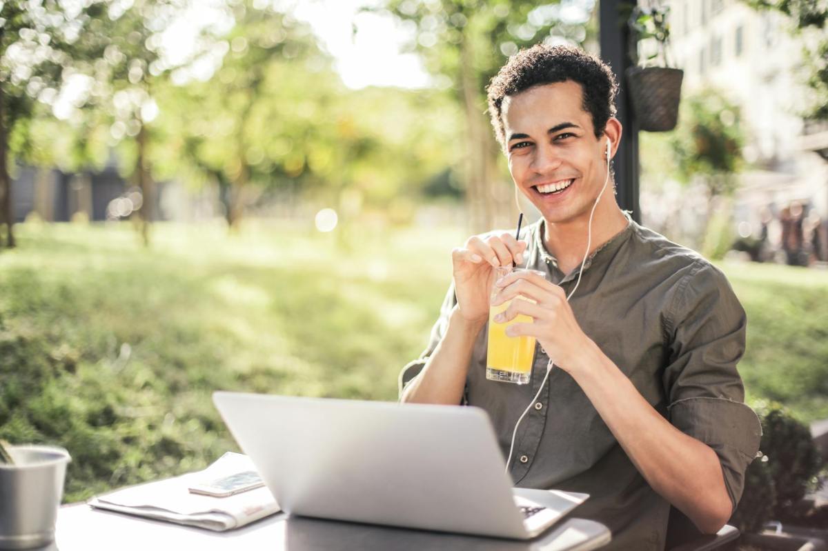 Young student siting outside drinking juice with an open laptop on the table.
