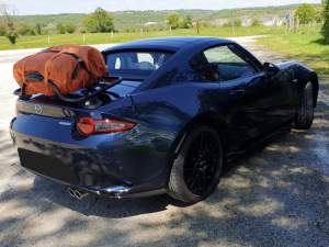 Mazda MX5 RF grise avec un porte-bagages Revo-Rack équipé d'un sac orange dans une aire de repos par une journée ensoleillée
