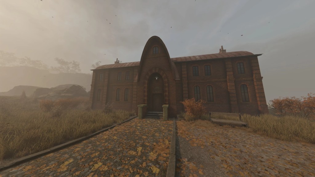 A large, brick building with a peaked roof and arched entrance, surrounded by overgrown grass and autumn foliage, under a cloudy sky.