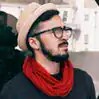 A bearded man rocking glasses, a sharp beige hat, and a bold red scarf over a dark shirt stands confidently outdoors, city buildings blurred in the background. He’s serving up boss-level style and attitude—unleashing urban energy for the world to see. - Boss Uncaged™ OmniMedia Inc.