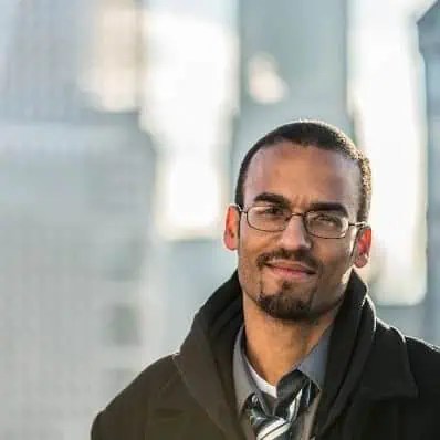 Donnie Bryant, sharp in glasses, goatee, dark coat, shirt, and tie, dominates the cityscape with urban buildings behind him. - Boss Uncaged™ OmniMedia Inc.