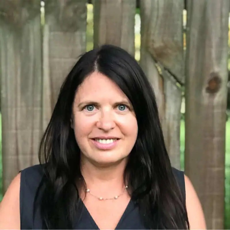 Confident woman with long dark hair in a sleek black sleeveless top, smiling powerfully in front of a rustic wooden fence. - Boss Uncaged™ OmniMedia Inc.