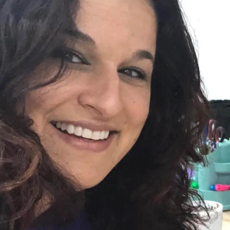 Confident woman with curly brown hair grins at the camera; dynamic workspace objects fade into a stylish, blurred background. - Boss Uncaged™ OmniMedia Inc.