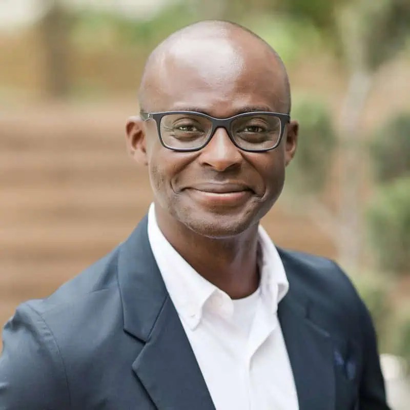 Confident man in glasses, dark blazer, and white shirt smiles at camera, exuding leadership against a blurred outdoor backdrop. - Boss Uncaged™ OmniMedia Inc.