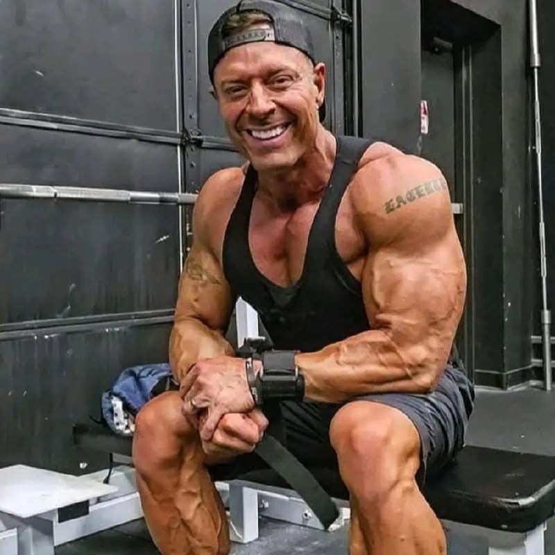 Confident, muscular man in black tank top and cap sits on gym bench, wrist straps on, smiling boldly into the camera. - Boss Uncaged™ OmniMedia Inc.