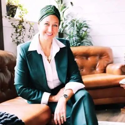 Confident woman in green suit and headscarf smiles on brown leather couch, surrounded by plants and white walls—boss vibes. - Boss Uncaged™ OmniMedia Inc.