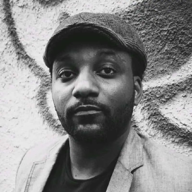 Powerful black and white portrait of Delray Nelson in a flat cap and jacket, posed confidently by a bold, graffiti-textured wall. - Boss Uncaged™ OmniMedia Inc.