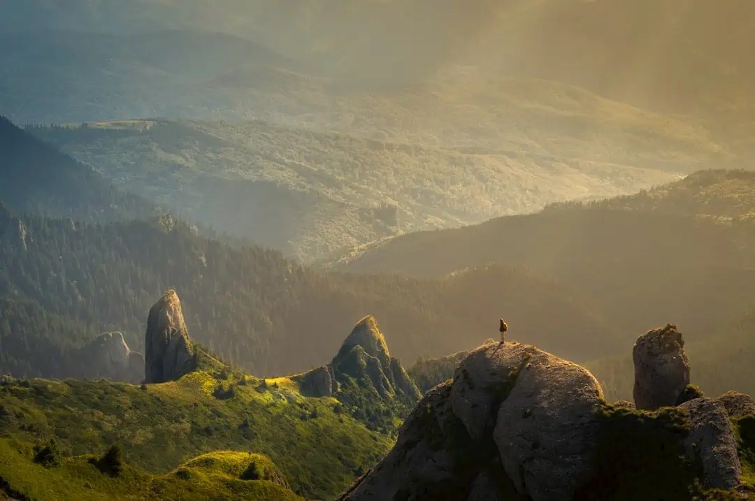 Dominate the summit: A trailblazer stands atop a rugged mountain peak, surveying vibrant valleys and rolling forested hills in golden light. - Boss Uncaged™ OmniMedia Inc.