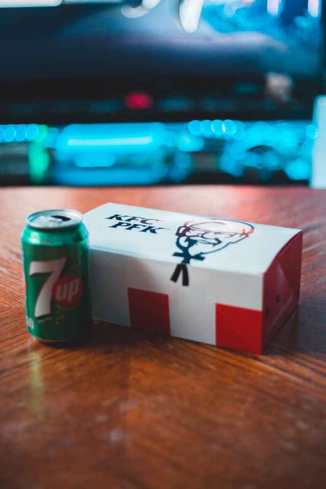 From above of metal can of beverage with cardboard box of food on wooden table near desktop computer glowing in apartment