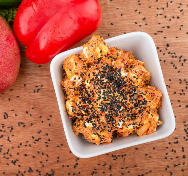 Top view of spicy cauliflower with black sesame in a square white bowl, surrounded by fresh vegetables.