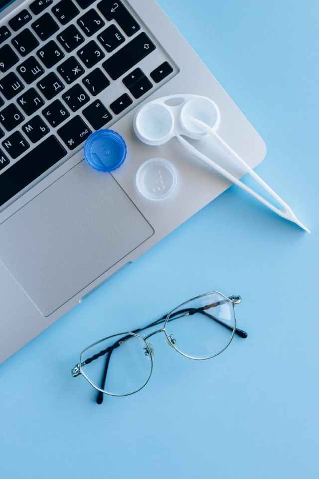 Flat lay of eyeglasses, contact lens case, and laptop on a blue surface.