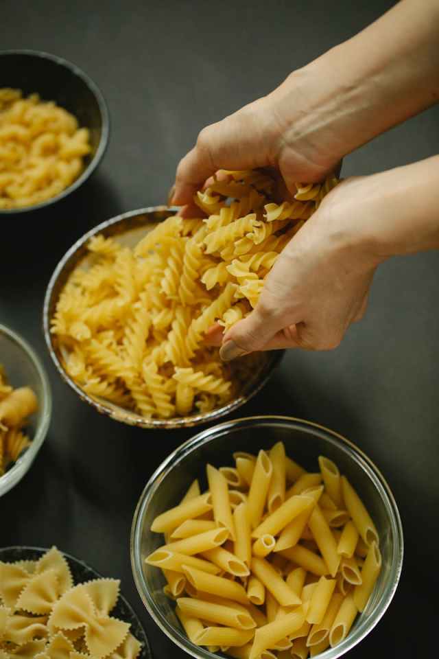 From above of crop anonymous female chef putting fusilli into bowl placed near macaroni and farfalle