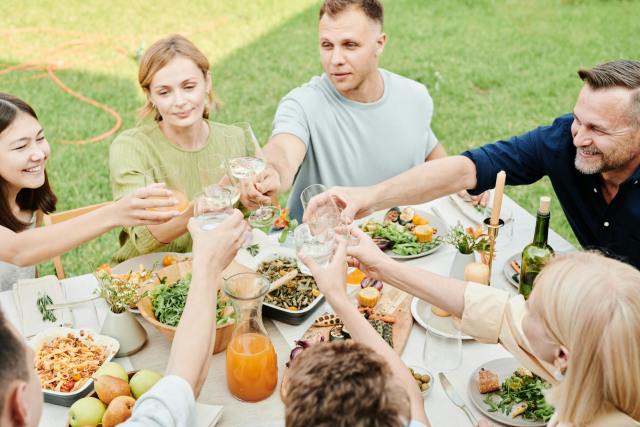 A joyful family with friends sharing a meal, toasting with drinks in a sunny outdoor setting.