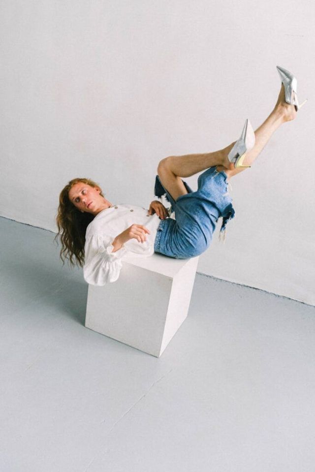 A model in denim and heels strikes a dynamic pose on a cube in a studio setting.