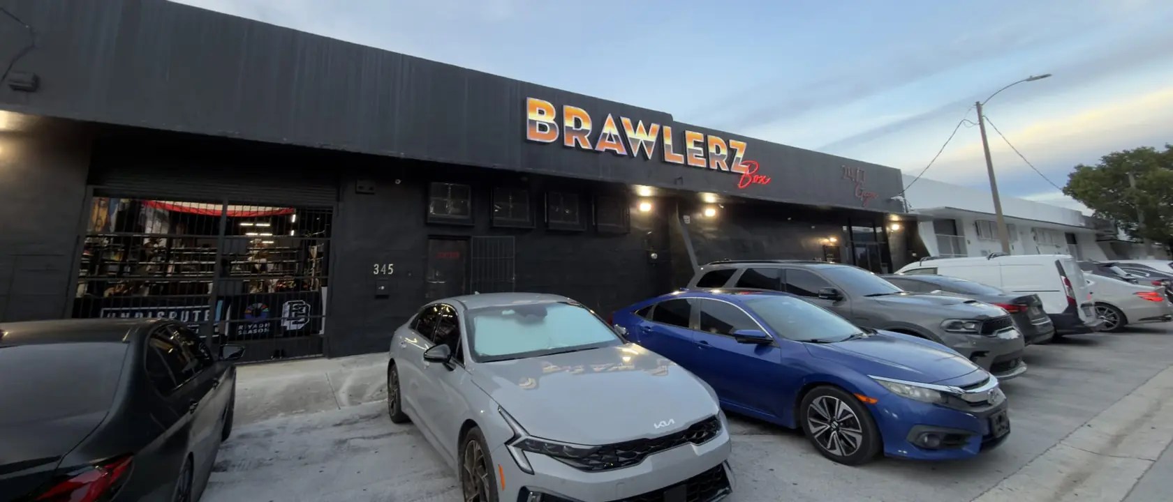 The black Brawlerz Box building at 345 NE 59th Street with its sunset sign lit at dusk and cars parked out front