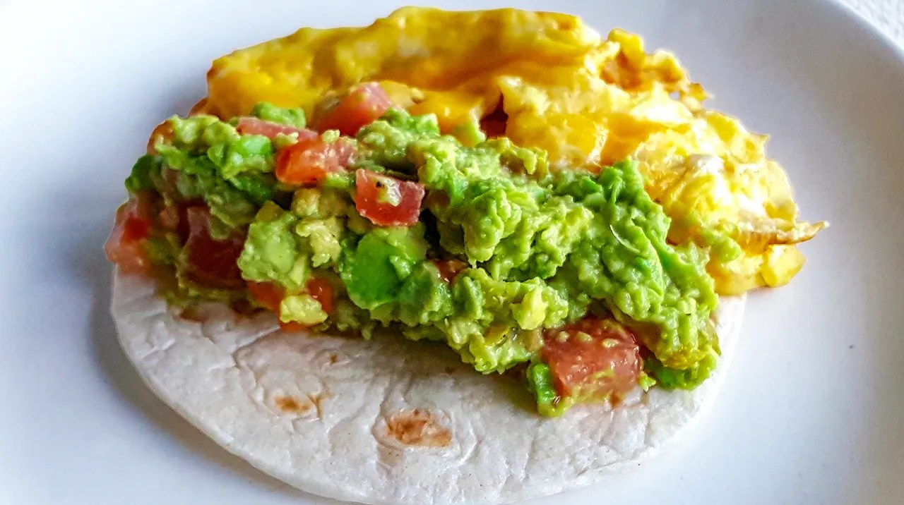 Scrambled eggs served with a tortilla topped with guacamole and diced tomatoes on a white plate