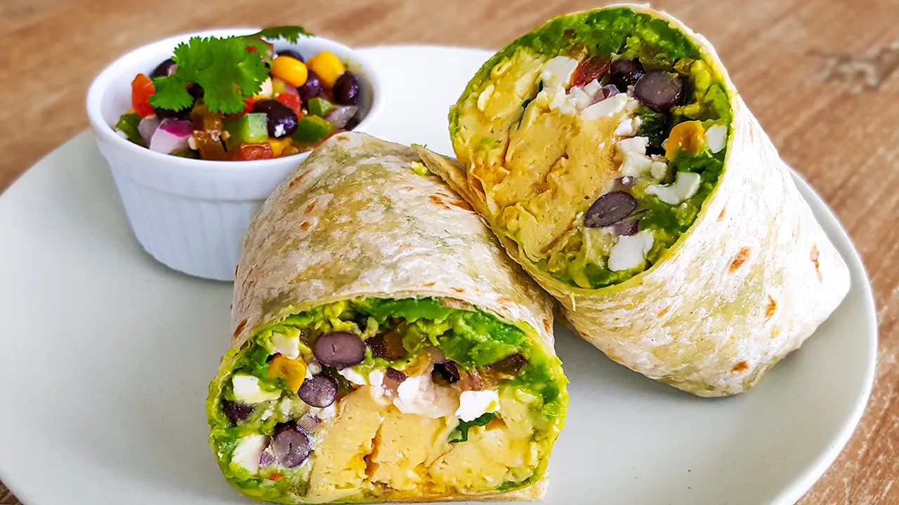 Two halves of a breakfast burrito filled with scrambled eggs, black beans, guacamole, and cheese on a white plate with a small bowl of corn and black bean salsa garnished with cilantro