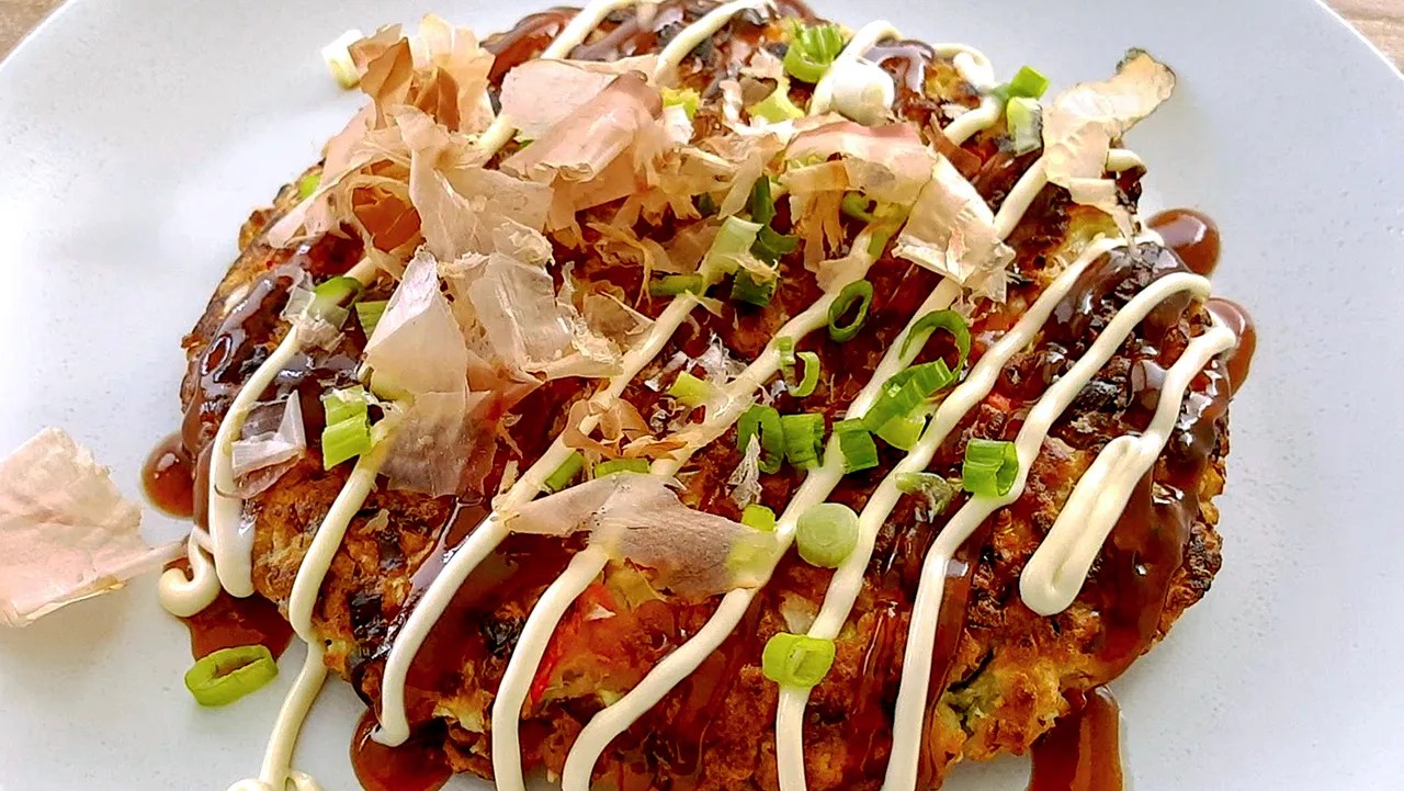 Okonomiyaki pancake topped with mayonnaise, okonomi sauce, green onions, and bonito flakes on a white plate