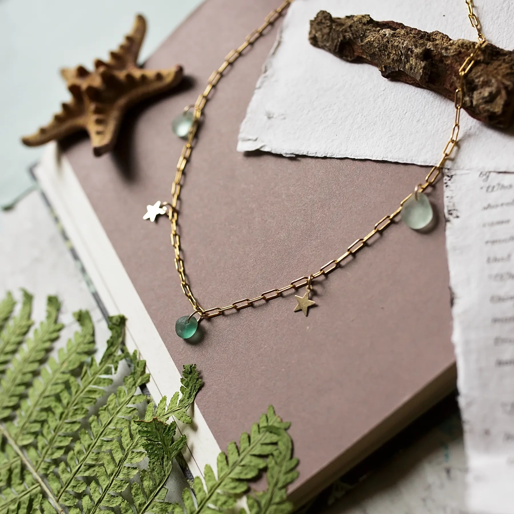 An elegant dangle Sea Glass necklace featuring green and teal sea glass charms and small 14Kgold filled star pendants, displayed on a soft, natural-themed background with greenery and torn paper textures. The design evokes a coastal and organic aesthetic.