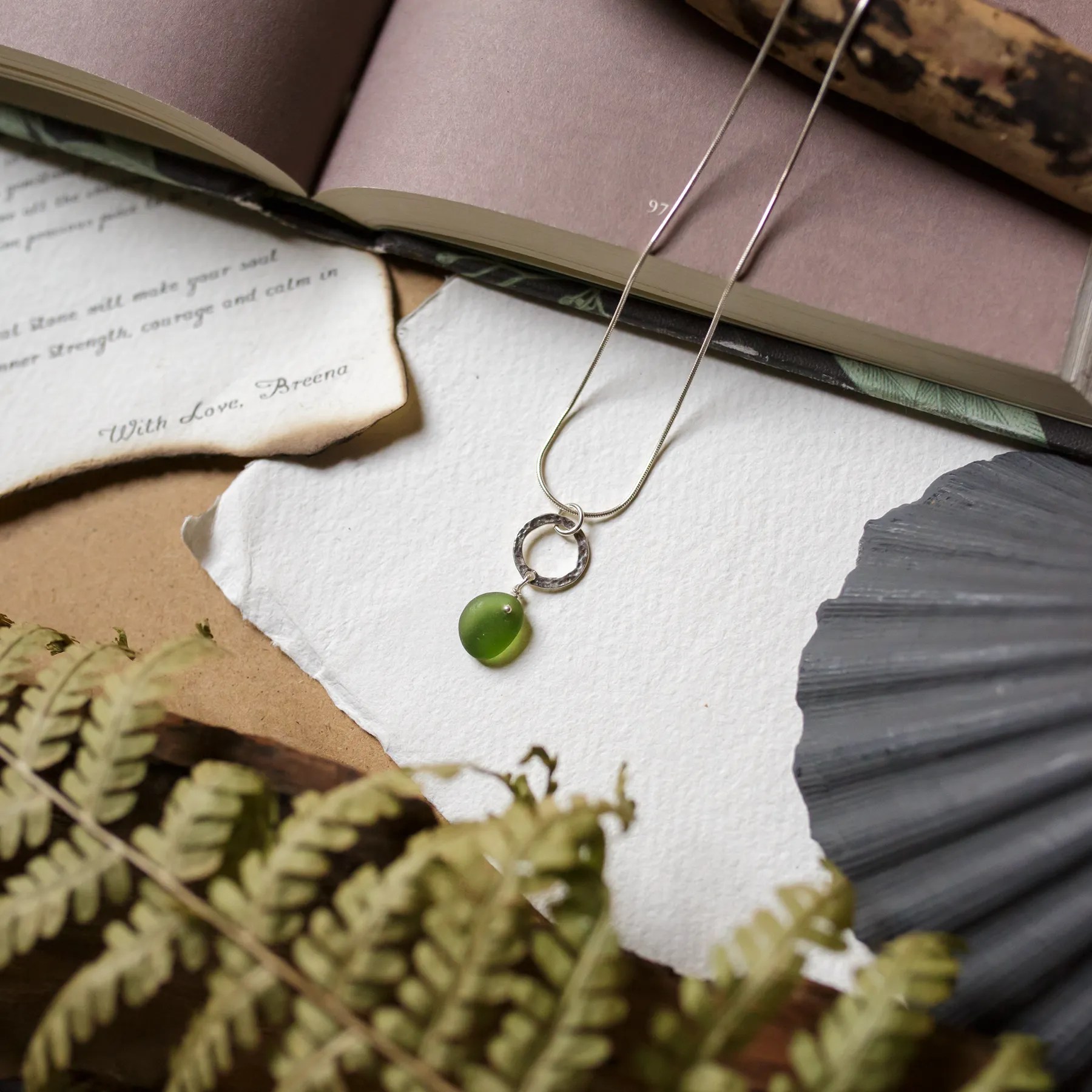 A delicate green Sea Glass necklace featuring green sea glass charm and hammered sterling silver ring, displayed on a soft, natural-themed background with greenery and torn paper textures. The design evokes a coastal and organic aesthetic.