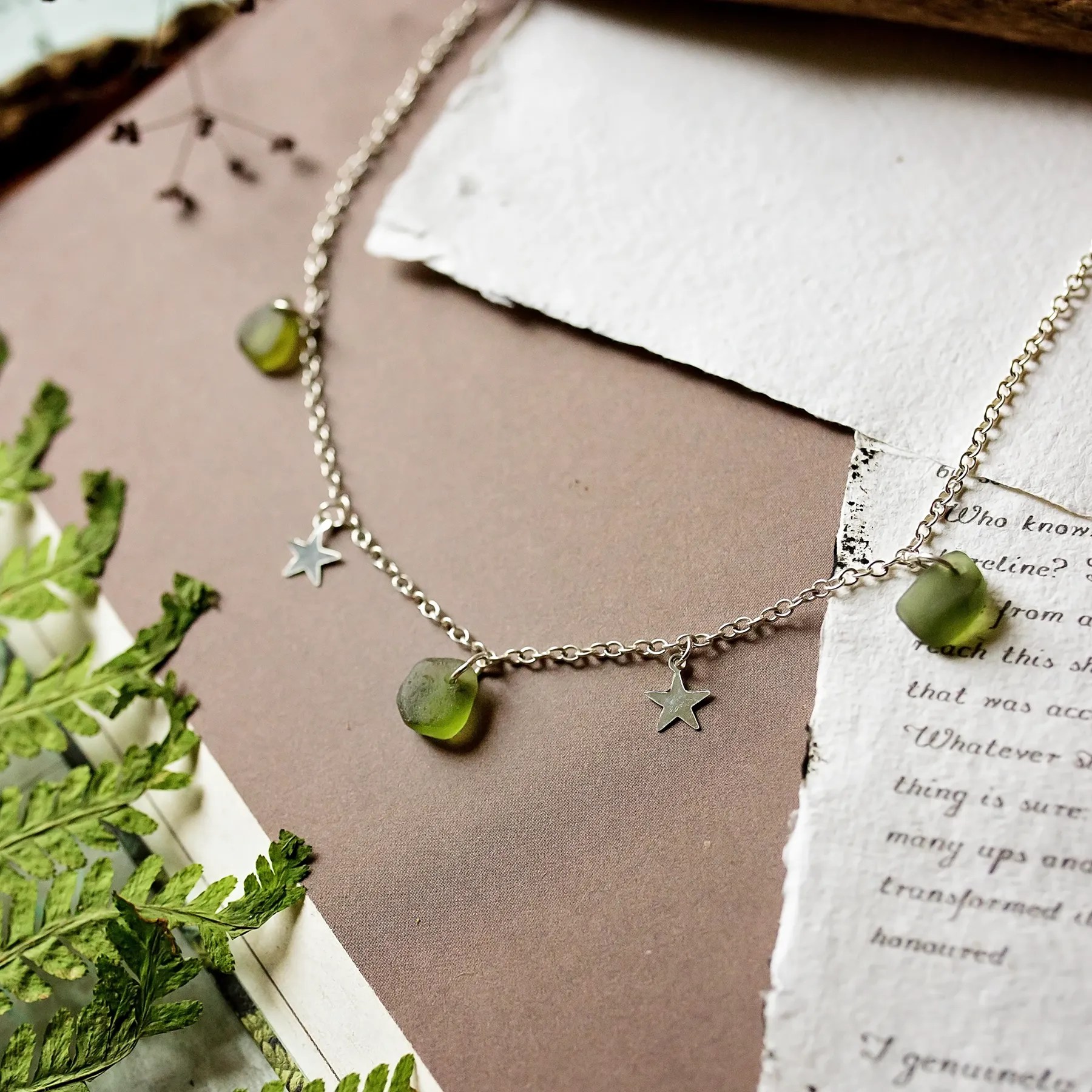 A delicate dangle Sea Glass necklace featuring green sea glass charms and small silver star pendants, displayed on a soft, natural-themed background with greenery and torn paper textures. The design evokes a coastal and organic aesthetic.