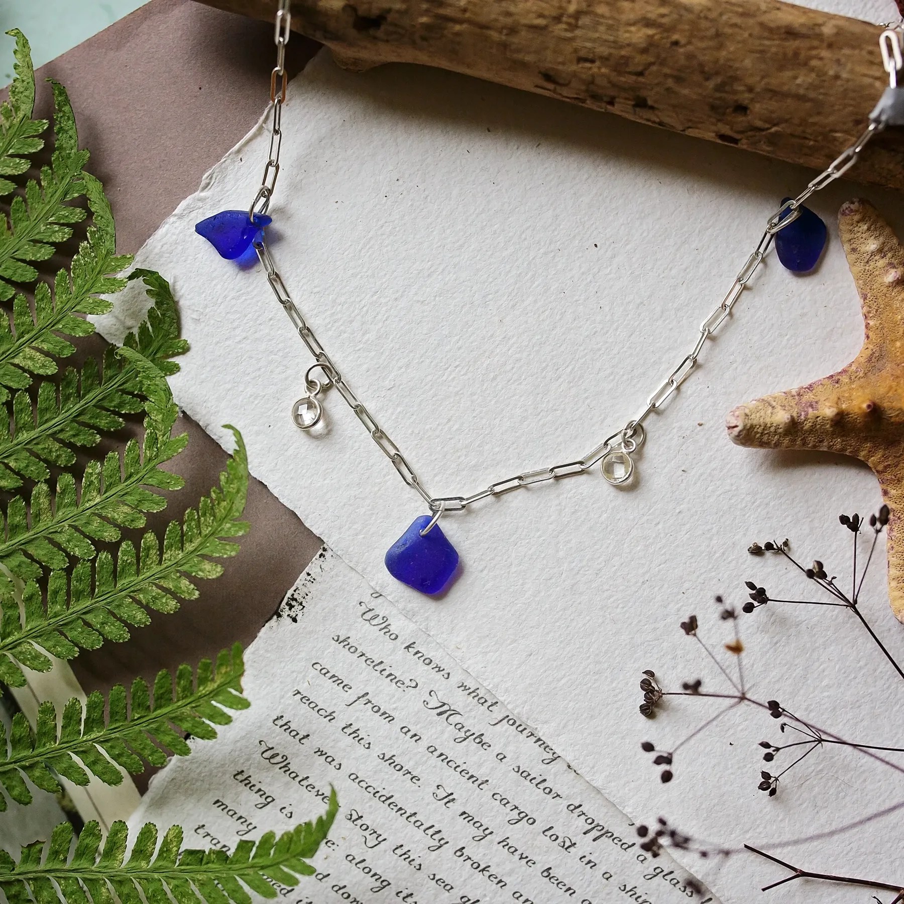A beautiful coastal inspired Sea Glass necklace featuring dark blue sea glass charms and small silver quartz pendants, displayed on a soft, natural-themed background with greenery and torn paper textures. The design evokes a coastal and organic aesthetic perfect for any ocean lover.