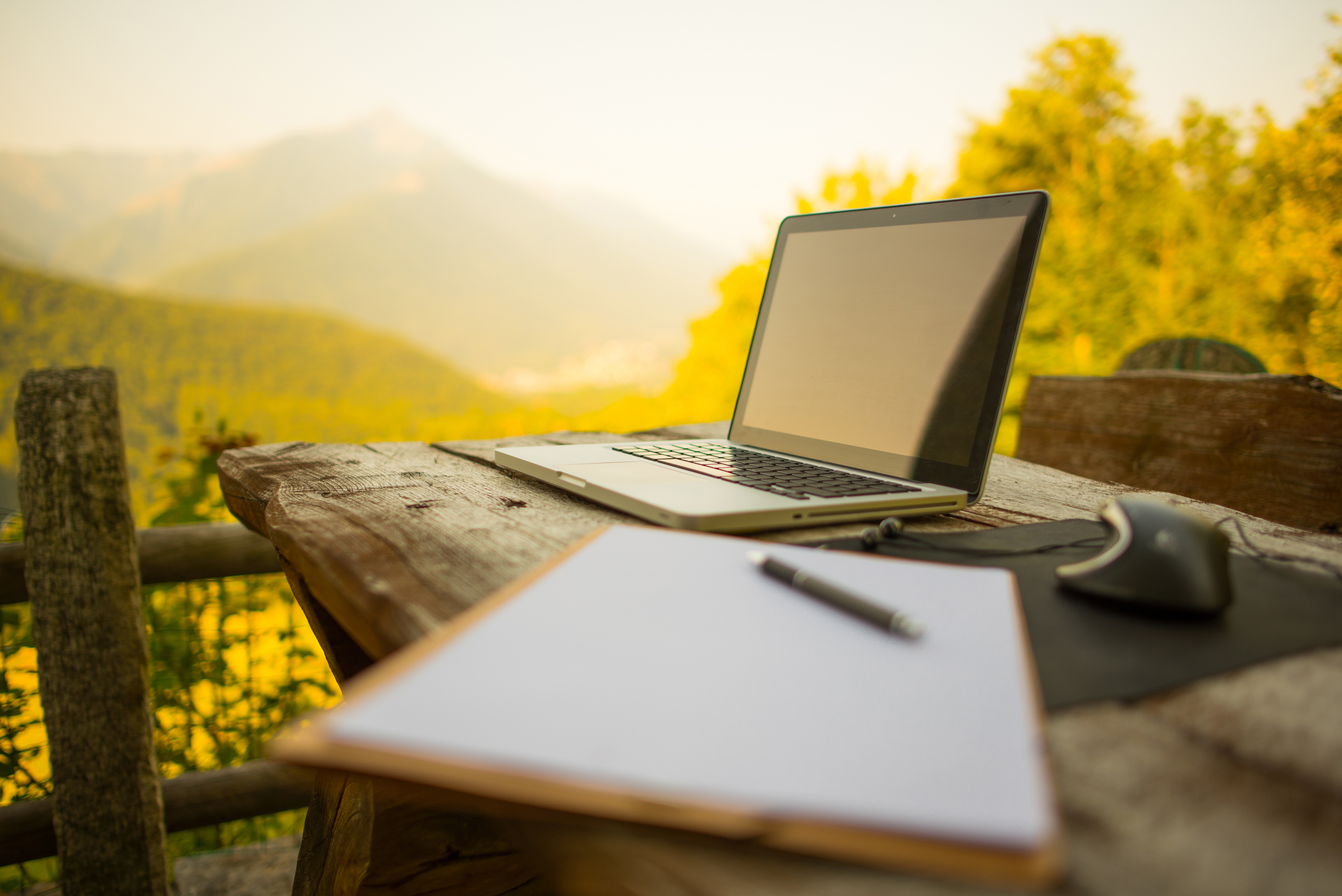Open laptop on a wooden table with notebook, pen, and mouse, set against a blurred mountain and forest background.