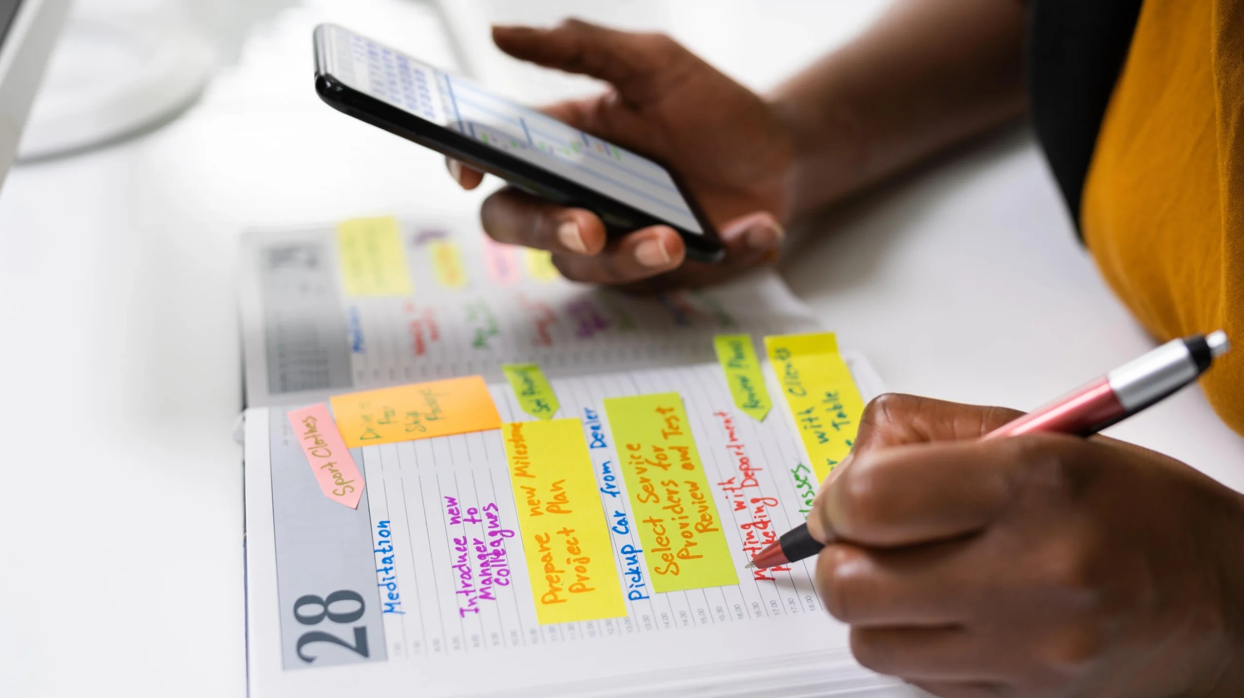 Person writing in a color-coded planner while referencing a smartphone schedule