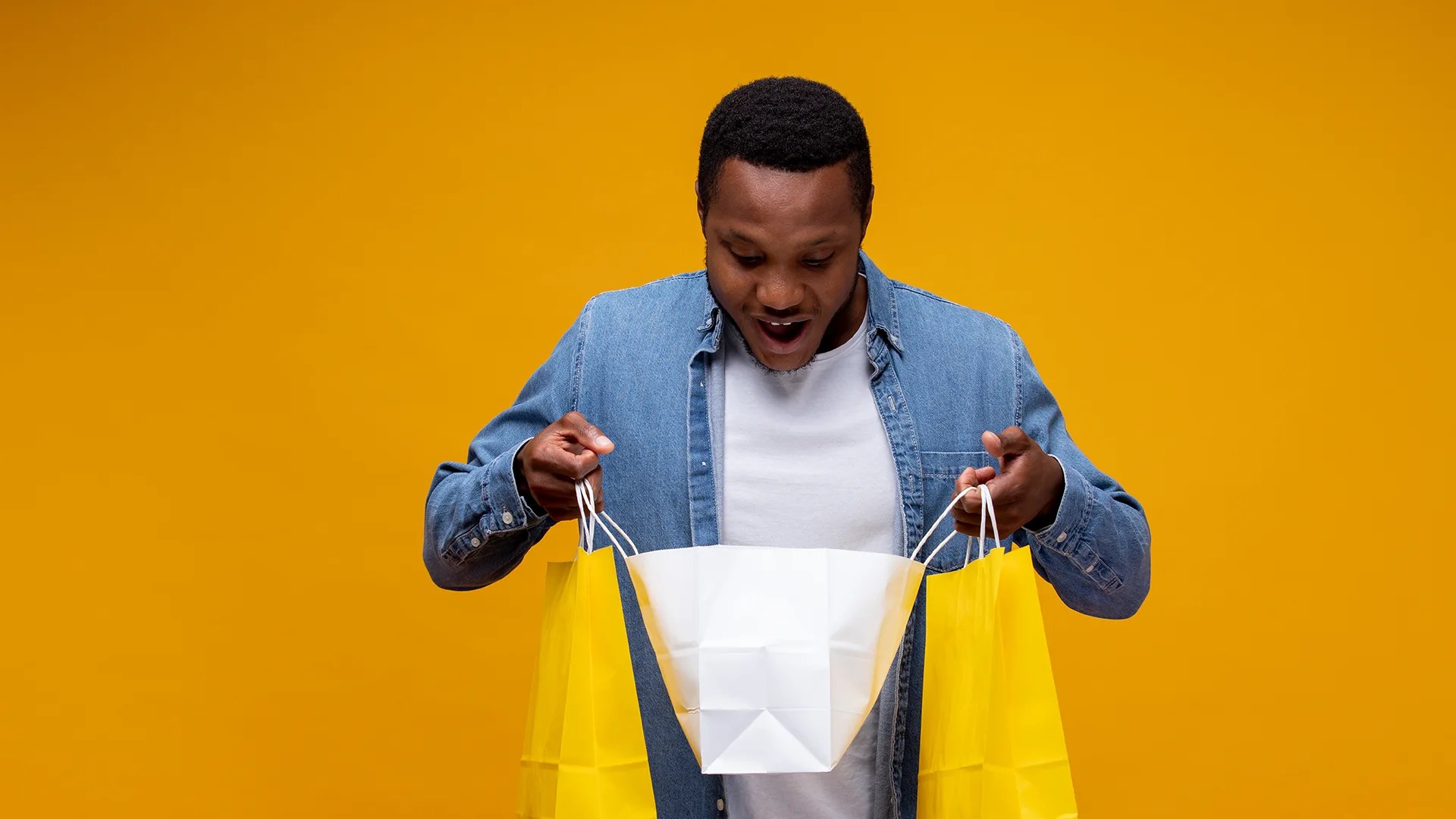 Excited man looking into shopping bags against a bright yellow background.