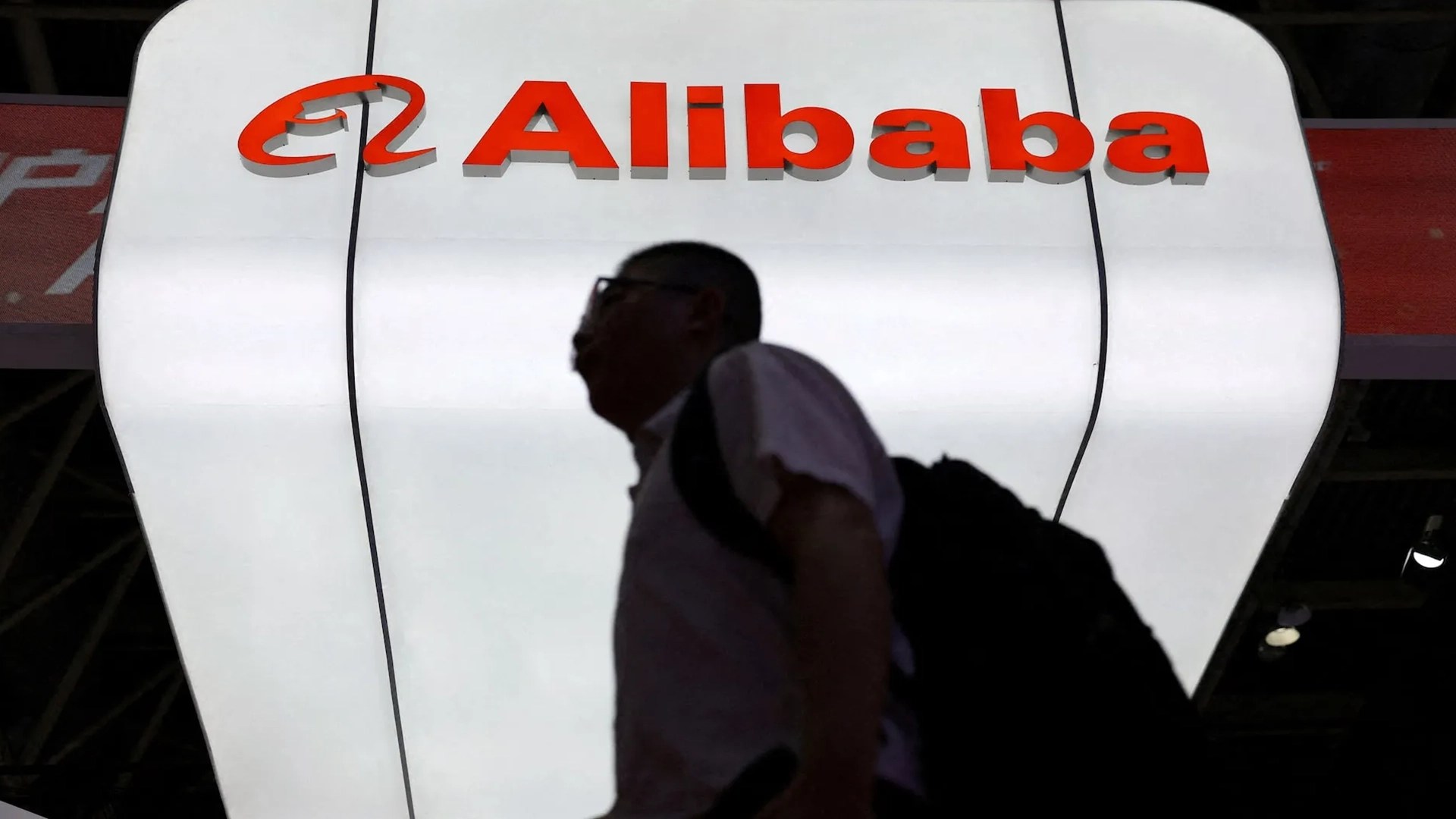 A large illuminated alibaba logo above a silhouetted person walking past at a technology exhibition
