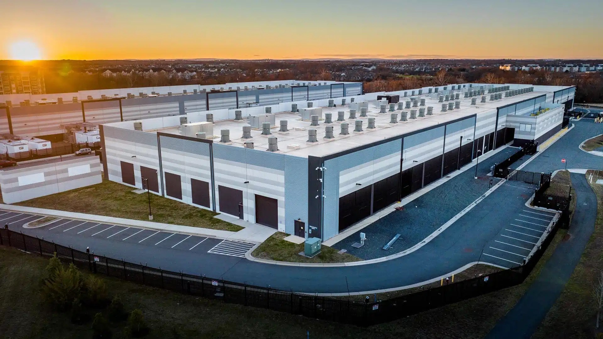 Aerial view of a large amazon data center complex at sunset showing long warehouse style buildings with rooftop cooling units surrounding roads and security fencing