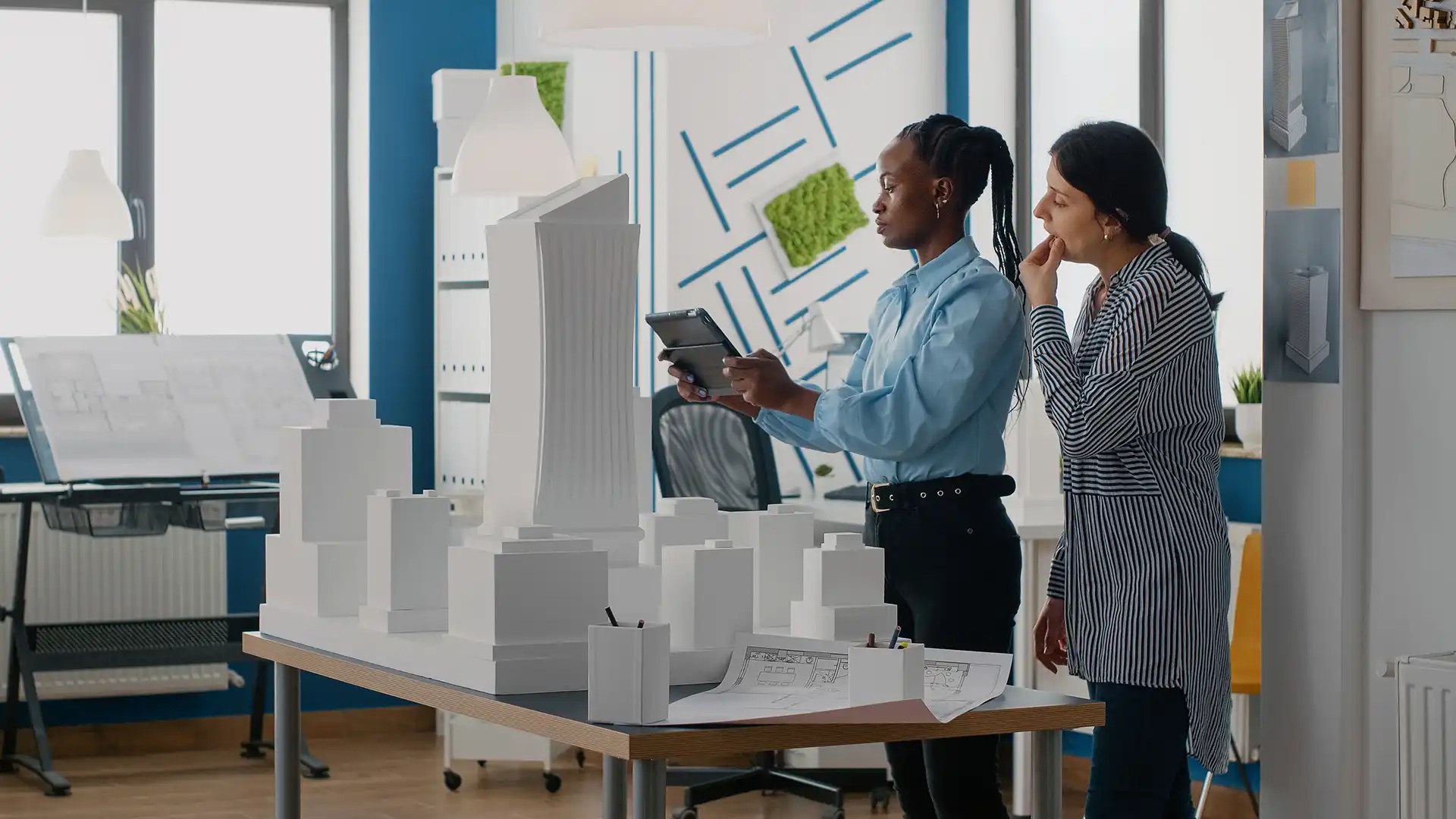 Two women in a bright design studio examine a tablet beside white architectural models on a table, discussing a project with scale models nearby and blueprints visible.