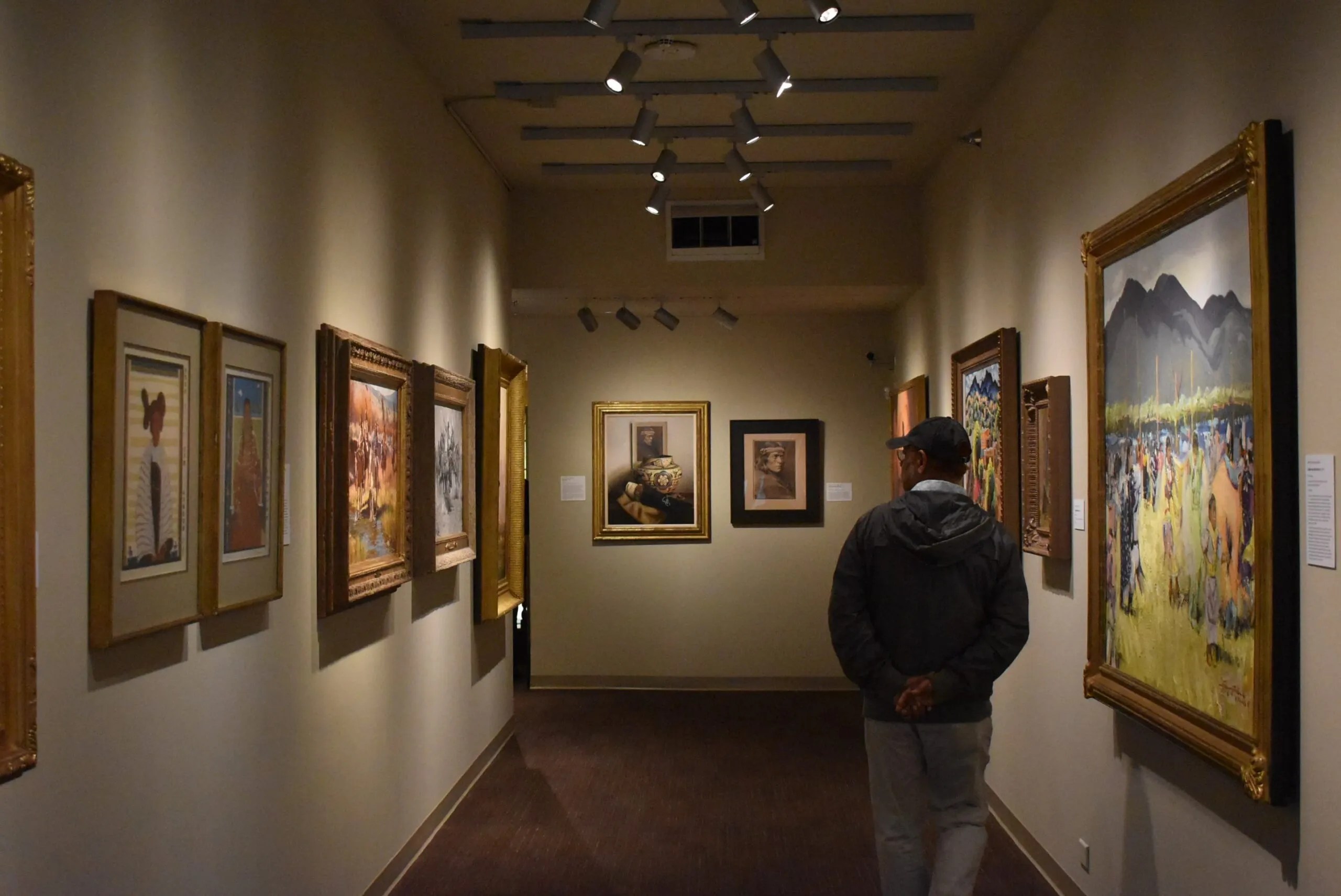 A person standing in an art gallery, observing various framed paintings displayed on the beige walls. The gallery has a warm and inviting atmosphere with soft lighting that highlights the artwork. The floor is carpeted, and there are track lights on the ceiling directed towards the paintings to enhance their visibility. The artworks feature diverse styles and subjects, including landscapes and still life.