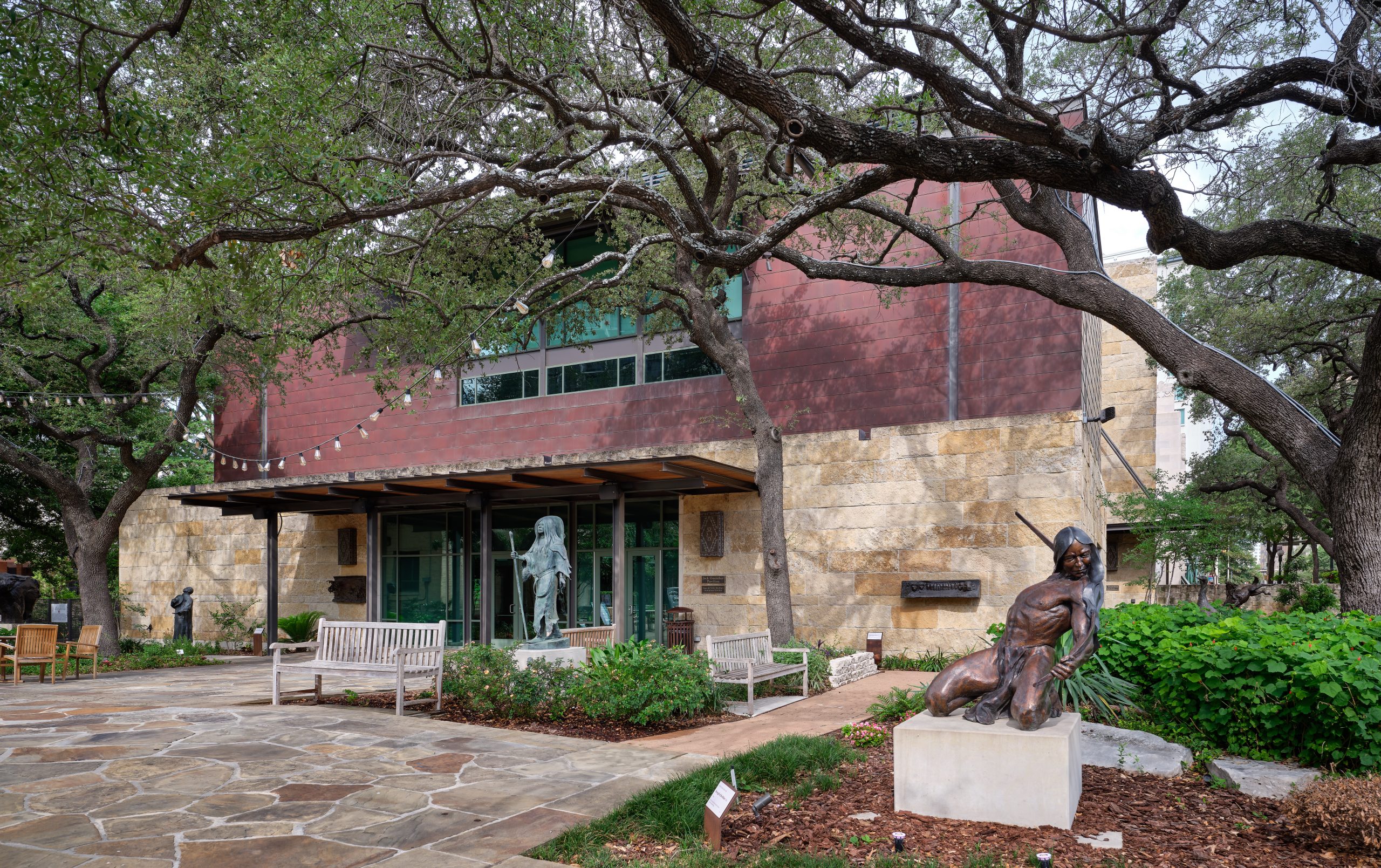 A building with natural stone walls and a large, sloping red roof, surrounded by mature trees. In front of the building, there is a paved area with benches and two bronze statues: one of a standing figure and another in a dynamic pose, possibly engaged in an activity like dancing or sports.
