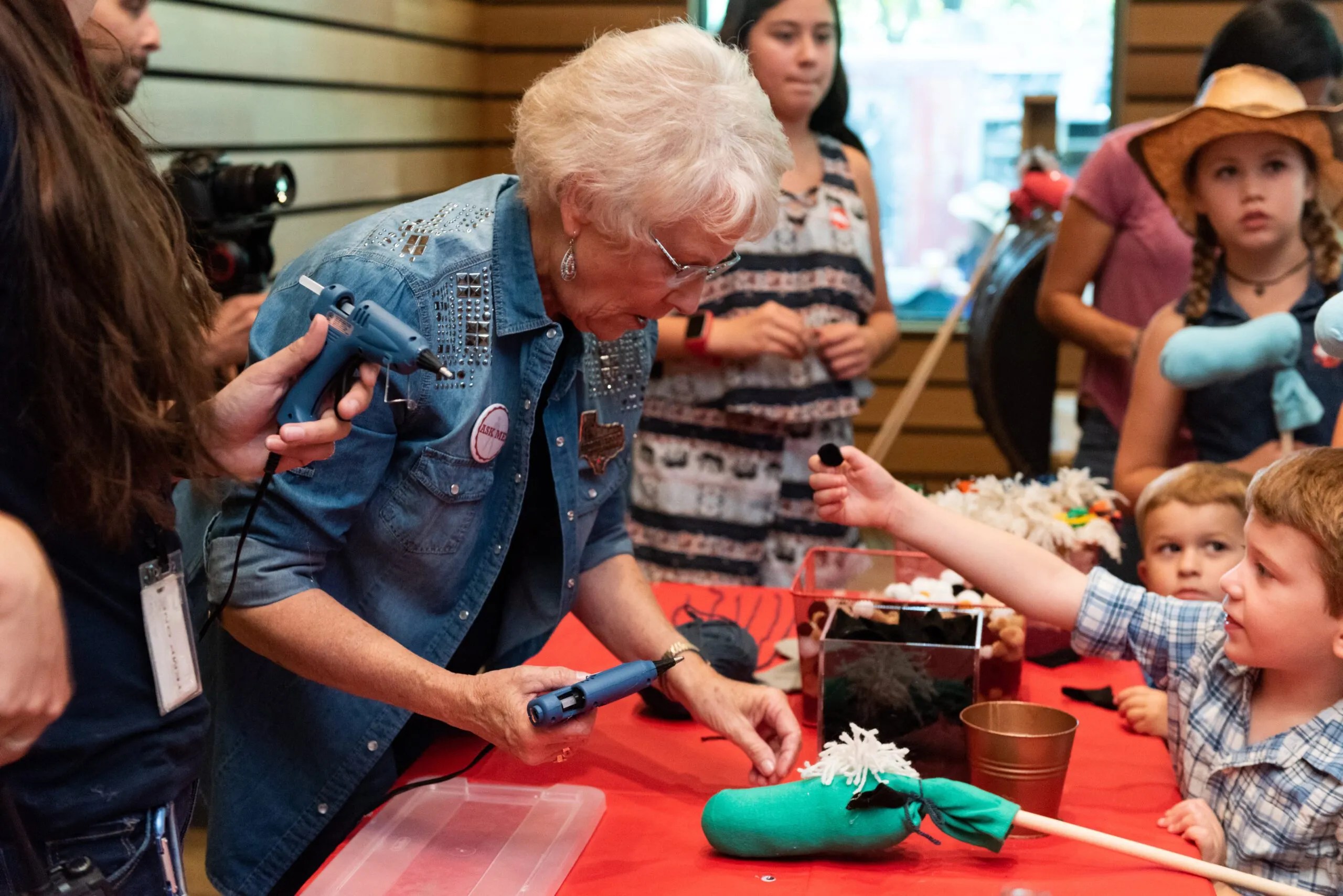 A older women helping hot glue eyes to a makeshift horse stick for a child. The child is holding the eye for her to glue on.