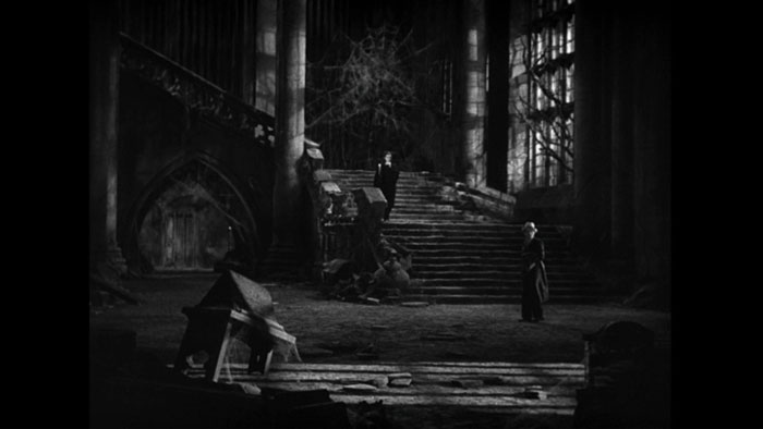 A black-and-white interior shot of a decrepit mansion foyer and once-majestic staircase: massive cobwebs, broken furniture, bare vines climbing through the windows. Dracula stands on a landing in the staircase; Harker, in a fedora and holding an overcoat over his arm, stands in the foyer.