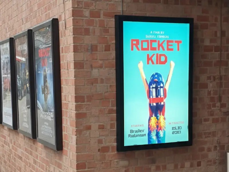 Illuminated black LED display case mounted on a brick wall, displaying a brightly lit poster for 'Rocket Kid', with three similar cases mounted beside it.