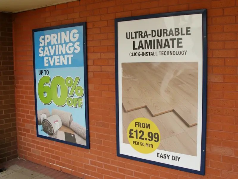 wo large Slimlock poster display cases mounted side-by-side on an exterior brick wall, featuring retail advertisements for a 'Spring Savings Event' and 'Ultra-Durable Laminate'.